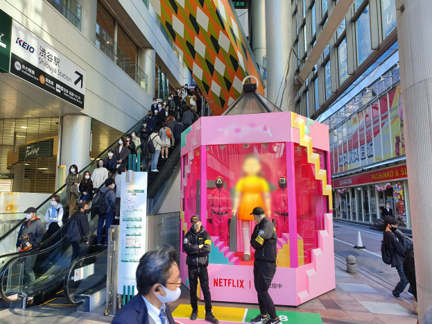 Eine Gruppe von Menschen steht vor einem Gebäude mit Glaswänden, Säulen und Texttafeln, einige tragen Masken, mit einer Rolltreppe im Blick; ein pinker Kasten in der Mitte zeigt Schaufensterpuppen und trägt "Netflix's new Netflix booth in Tokyo."