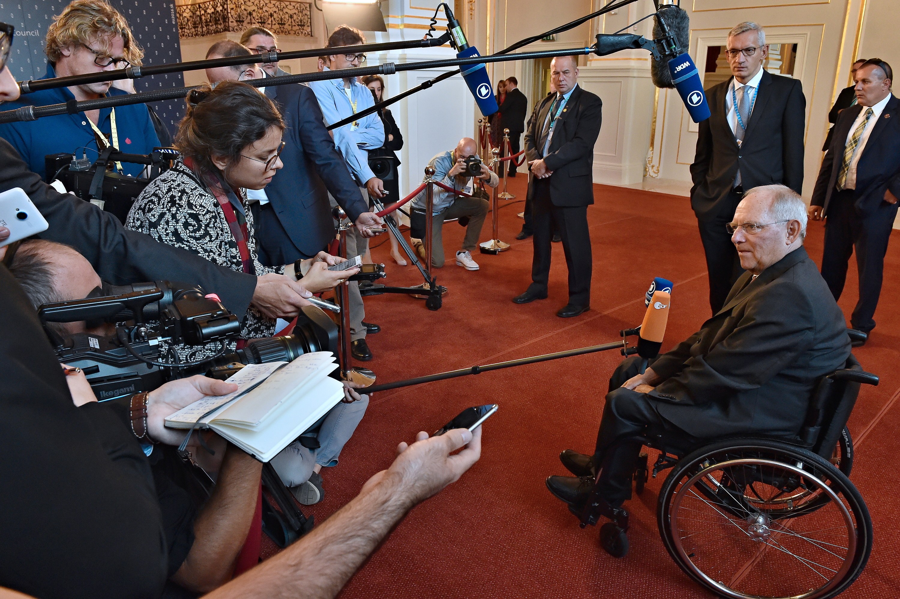 Ein Mann im Rollstuhl spricht zu einer Gruppe von Reportern mit Kameras, Mikrofonen und Handys, mit einem Banner, Scheinwerfern und einer Wand im Hintergrund.