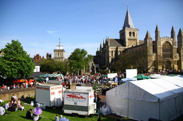 Historische Gebäude mit Bäumen davor, zusätzliche Gebäude und ein Turm im Hintergrund, Menschen auf einem Weg mit Geschäften daneben und andere auf der nahen Wiese sitzend.