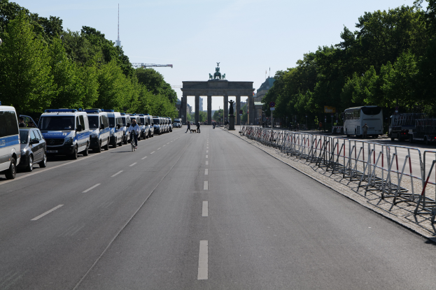 Eine Reihe von Polizeiwagen, die auf einer Straße vor dem Brandenburger Tor in Berlin, Deutschland, geparkt sind, mit Fahrradfahrern, Fußgängern, Barrieren, Bäumen und einem Bogen mit Statuen im Hintergrund.
