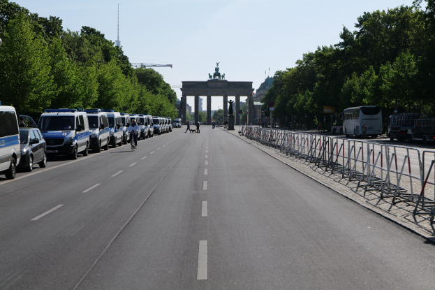 Lange Reihe von Polizeiwagen am Straßenrand vor dem Brandenburger Tor in Berlin, Deutschland, mit Menschen auf Fahrrädern und Absperrungen auf der Straße, Bäumen an den Seiten und einem Bogen mit Statuen im Hintergrund.