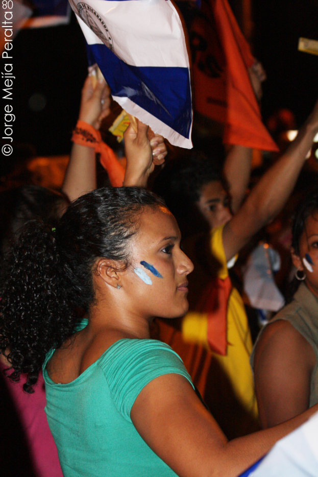 Eine Frau in einem grünen T-Shirt steht auf einer Demo und hält ein weißes Schild hoch, mit vielen anderen Menschen dahinter.