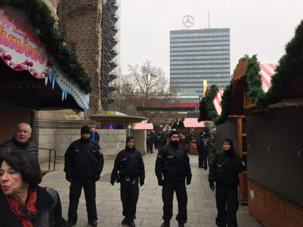 Eine Gruppe von Polizisten in schwarzen Uniformen steht vor einem festlich geschmückten Weihnachtsmarkt in Berlin, Deutschland, mit Gebäuden, Bäumen und einem klaren blauen Himmel im Hintergrund und einer Tafel mit Text auf der linken Seite des Bildes.
