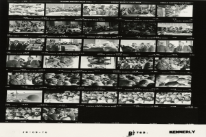 Schwarzes und weißes Foto einer vielfältigen Menschenmenge, die fröhlich in Richtung eines Fernsehbildschirms schaut, mit Text am unteren Rand.