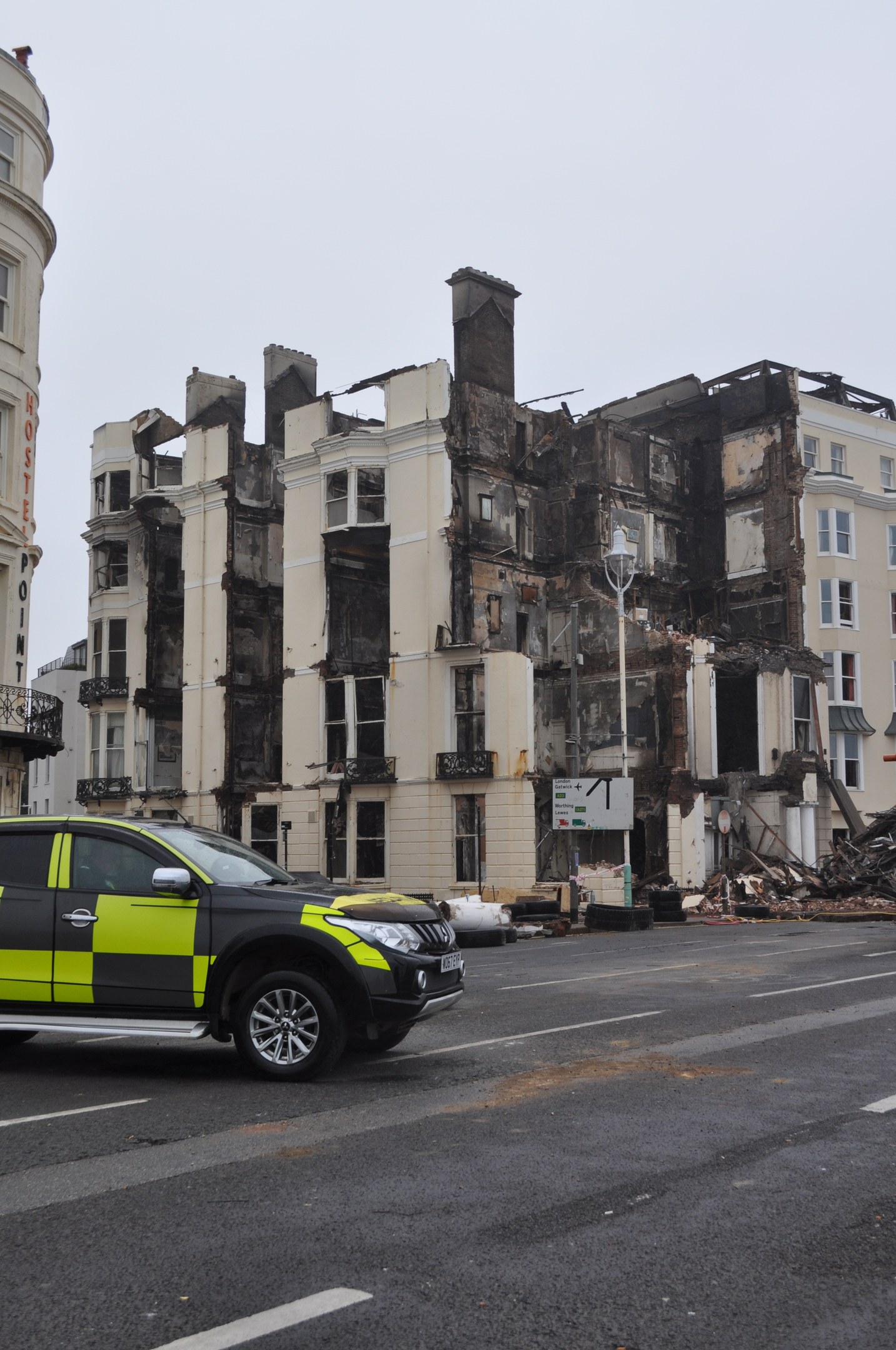 Ein Polizeiwagen steht vor einem schwer beschädigten Gebäude mit zersplitterten Fenstern und verstreuter Trümmer, mit anderen Gebäuden und dem Himmel im Hintergrund.