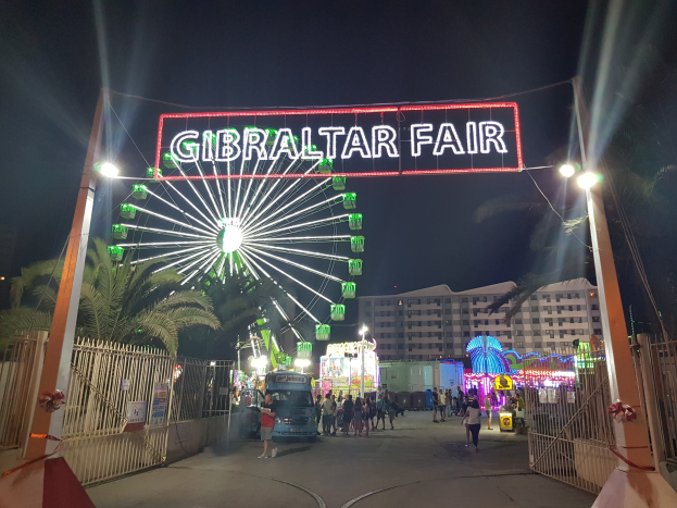 Eingang zur Gibraltar-Messe nachts mit beleuchtetem Riesenrad, beleuchteter Namensanzeige, Bäumen und Gebäuden im Hintergrund bei dunklem Himmel.