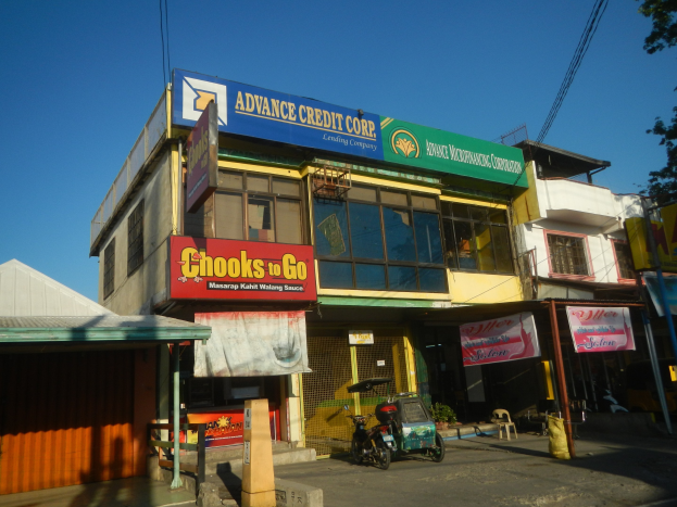 Gebäude mit "Advance Credit Corp"-Schriftzug, umgeben von Fahrzeugen, Werbetafeln, Strommasten, Kabeln, Bäumen und einem klaren blauen Himmel.
