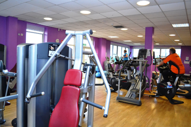 Ein Fitnessstudio mit verschiedenen Geräten und einigen trainierenden Personen, mit einer Decke und Lampen an der Oberseite.