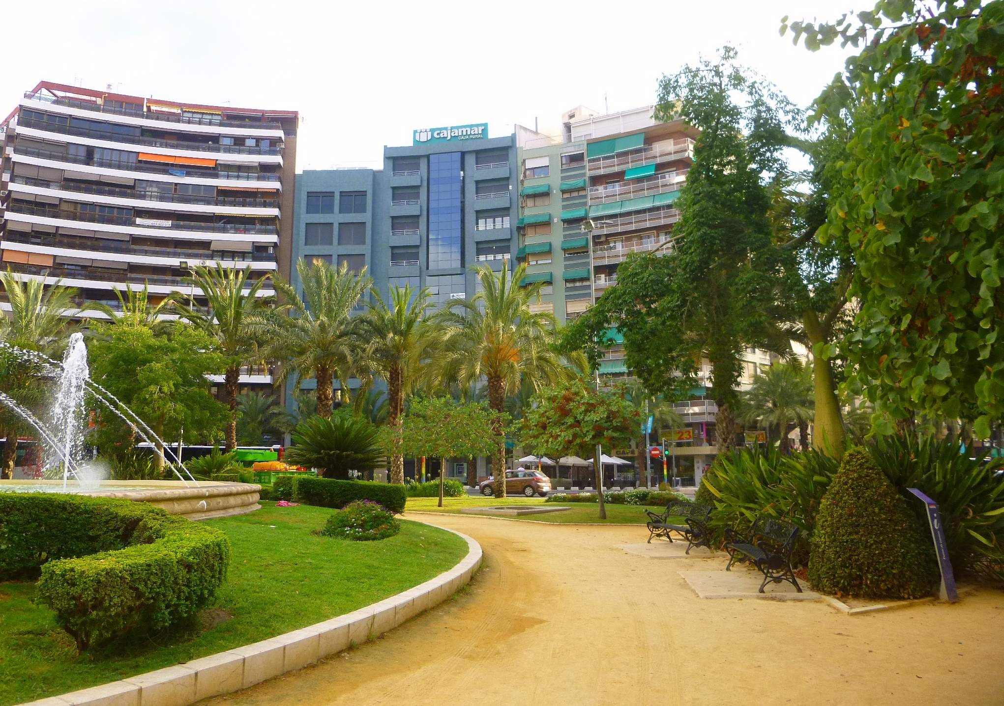 Ein Park mit einem zentralen Brunnen, umgeben von Grünflächen einschließlich Rasen, Pflanzen und Bäumen, mit Bänken, Laternenmasten und Straßenfahrzeugen sowie hohen Gebäuden und einem klaren blauen Himmel im Hintergrund.
