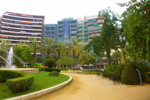 Ein Park mit einem zentralen Brunnen, umgeben von Grünflächen einschließlich Rasen, Pflanzen und Bäumen, mit Bänken, Laternenmasten und Straßenfahrzeugen sowie hohen Gebäuden und einem klaren blauen Himmel im Hintergrund.