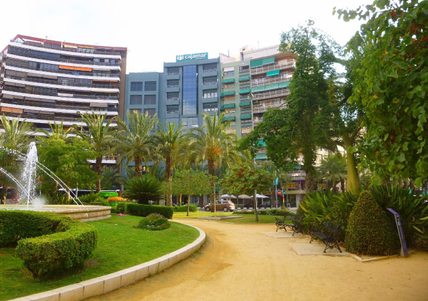 Ein Park mit einem zentralen Brunnen, umgeben von Grünflächen einschließlich Rasen, Pflanzen und Bäumen, mit Bänken, Laternenmasten und Straßenfahrzeugen sowie hohen Gebäuden und einem klaren blauen Himmel im Hintergrund.