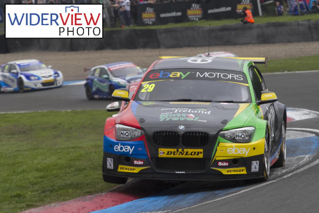 Rennwagen auf einer Strecke mit einer Gruppe von Menschen, Gras und Werbetafeln im Hintergrund und einem Wasserzeichen auf dem Bild.
