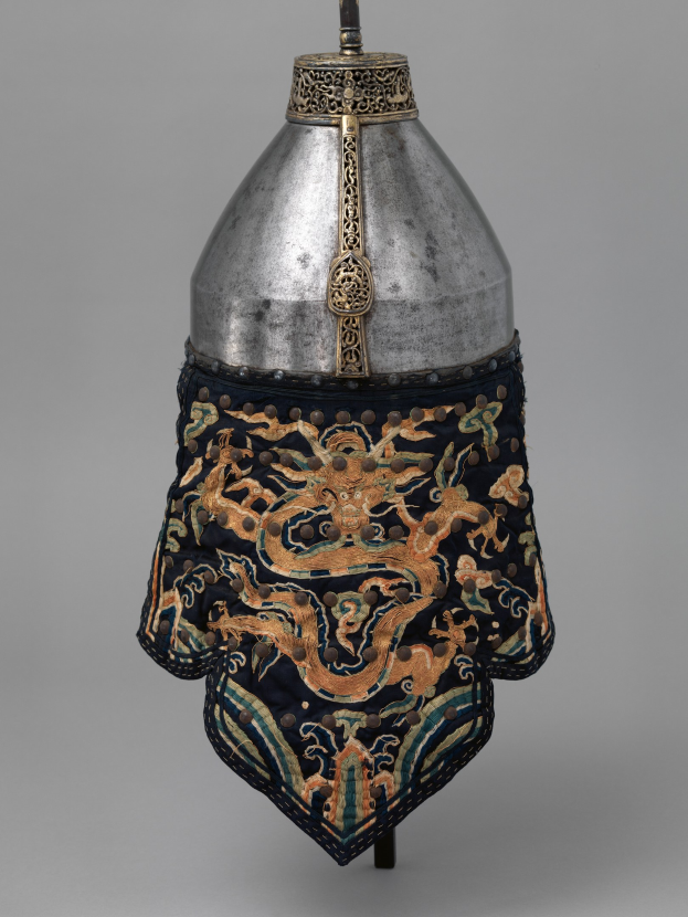 Metallhelm mit Drachendesign vor grauem Hintergrund.