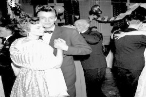 Ein Schwarz-Weiß-Foto von einem Mann und einer Frau, die auf einer Party tanzen, umgeben von lachenden Zuschauern und Ballons sowie Dekorationen im Hintergrund.