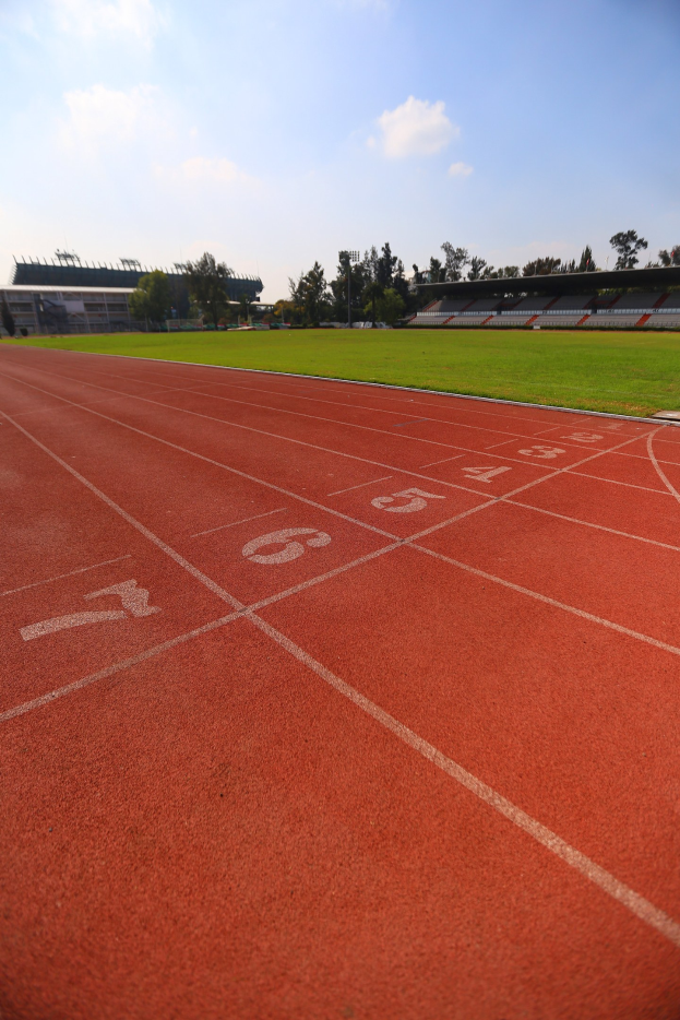 Laufbahn mit Bahnnummern, umgeben von grünem Gras und Bäumen, mit einem Stadion und einem klaren blauen Himmel im Hintergrund.