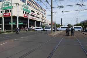 Eine Gruppe von Polizisten steht an der Straßseite neben einer Tram, mit Fahrzeugen, Strommasten, Gebäuden mit Namensschildern, Bäumen und einem bewölktem Himmel im Hintergrund.