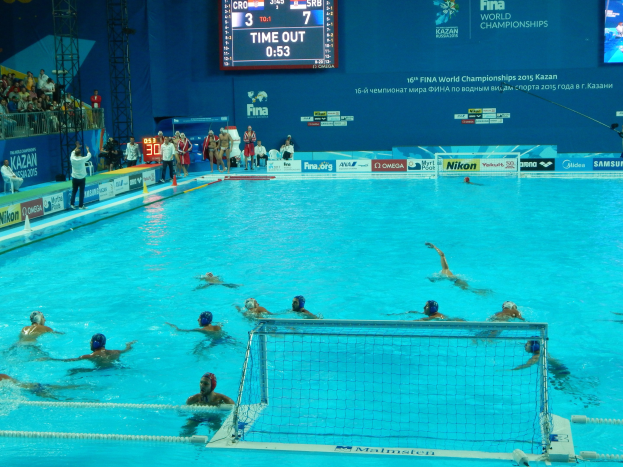 Gruppe von Athleten beim Wasserballspielen in einem Pool während der 2015 FINA-Weltmeisterschaften in Kazan, mit Zuschauern und Eventwerbung im Hintergrund.