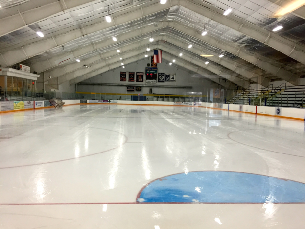 Großes Hallen-Eisstadion mit Deckenbeleuchtung, Werbetafeln, Treppen mit Geländern und wenigen Personen im Hintergrund.