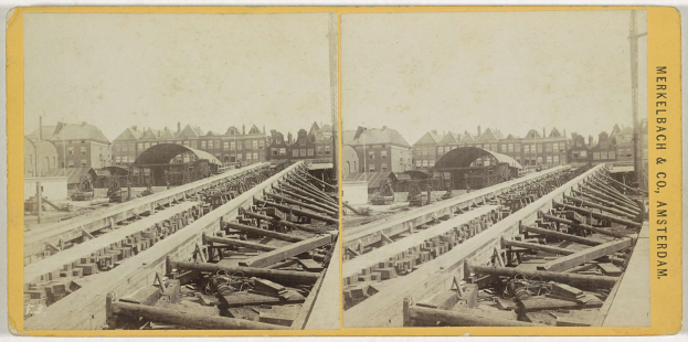 Schwarz-weiß-Foto einer Zugschiene mit Gebäuden im Hintergrund, Text "Merkelbach & Co, Co. Amsterdam" auf der rechten Seite und verstreute hölzerne Gegenstände im Vordergrund.