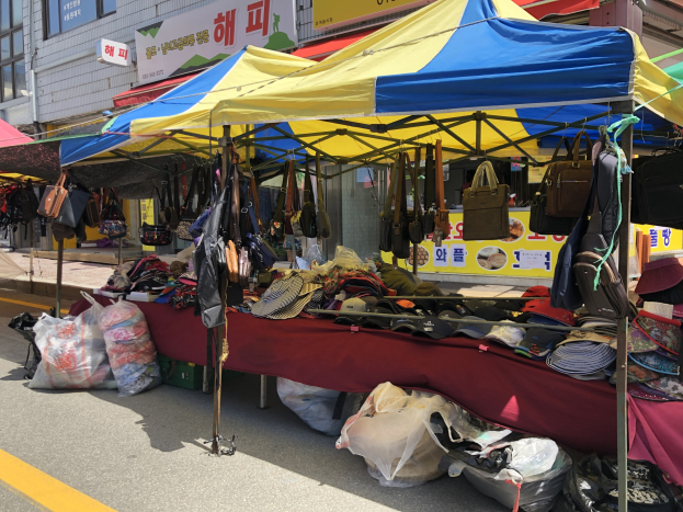 Ein Markt mit Zelten, die Taschen, Hüte und andere Gegenstände präsentieren, vor einem Hintergrund aus Gebäuden mit Fenstern und Texttafeln.