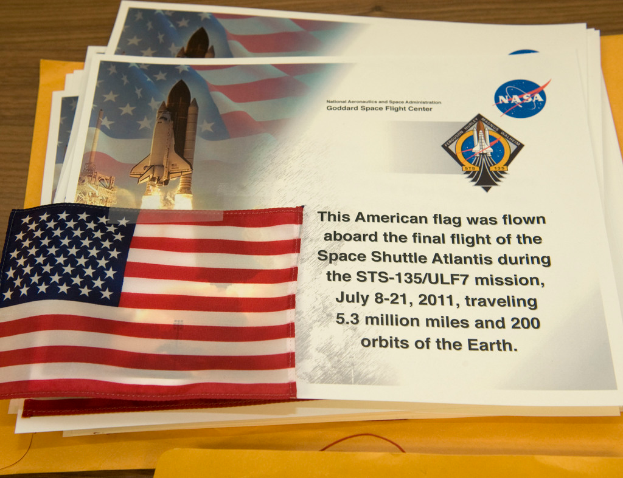 Ein zentrales Display zeigt Plakate mit Text, einer Flagge, einer Rakete und verschiedenen Logos, mit einem Tisch unten und einem Boden oben im Bild.