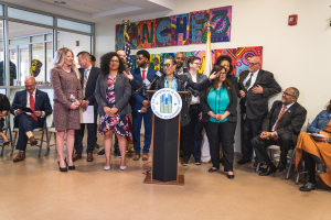 Gruppe von Menschen vor einem Rednerpult mit Mikrofon und einer Tafel mit der Aufschrift "Sancho City Councilman Joins City Council", mit sitzenden und stehenden Gästen, Flaggen, Feuerlöscher und Deckenlampen im Hintergrund.