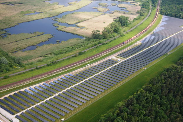 Luftaufnahme einer Solaranlage mit Solarpanelen auf einem Feld, umgeben von Bäumen, Gras, Pflanzen und Wasser, mit einem Zug auf einer nahen Bahnstrecke.