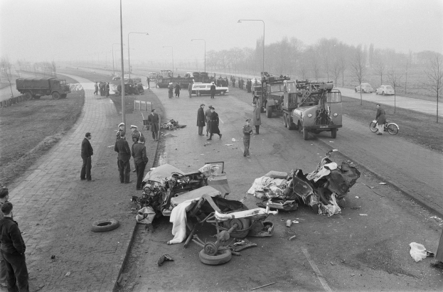 Schwarz-weißes Bild eines Autounfalls am Straßenrand mit mehreren Fahrzeugen und einer Gruppe von Menschen in der Nähe, Straßenlaternen, Bäumen und Himmel im Hintergrund.
