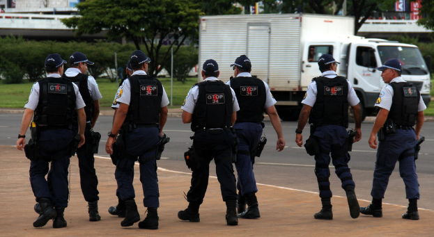 Eine Gruppe von Beamten in Uniform, einschließlich Mützen, Hemden, kugelsicheren Westen, Uhren, Hosen und Schuhen, die auf einer Straße mit einem geparkten Lastwagen auf der rechten Seite neben einem Zaun, Gebäuden, Bäumen, Pflanzen, Polen und Bannern im Hintergrund und einer Brücke im oberen rechten Eck gehen.