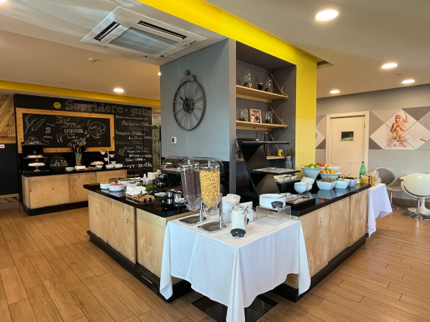 Ein Restaurantinnenraum mit Tischen und Stühlen, Essensbehältern, einer Tafel mit Text, einer Wanduhr, Deckenleuchten, einer Klimaanlage und einer Tür im Hintergrund, das sich im Hôtel de Ville in Paris befindet.