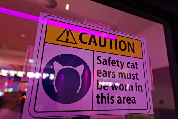 Hellgelbes Warnschild in einem Ladfenster mit der Aufschrift "Vorsicht: Sicherheitskatzenohren müssen in diesem Bereich getragen werden." vor einem unscharfen Hintergrund.