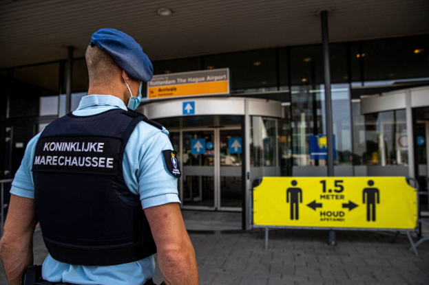 Polizeibeamter in blauer Uniform und Maske vor einem Glasgebäude mit mehreren Informationsschildern in der Nähe.