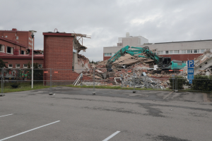 Großes Gebäude auf einem Parkplatz mit einem Bagger, Metallstangen, Zaun, Schild, Pflanzen, Gras, Bäumen, benachbarten Gebäuden und einem bewölkten Himmel abgebrochen.