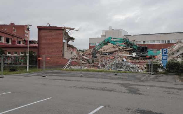 Großes Gebäude auf einem Parkplatz mit einem Bagger, Metallstangen, Zaun, Schild, Pflanzen, Gras, Bäumen, benachbarten Gebäuden und einem bewölkten Himmel abgebrochen.