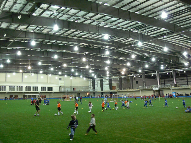 Eine Hallenfußballhalle mit einer Gruppe von Menschen, die mit einem Fußball auf dem Boden spielen, Torpfosten und Beleuchtung auf dem Dach.