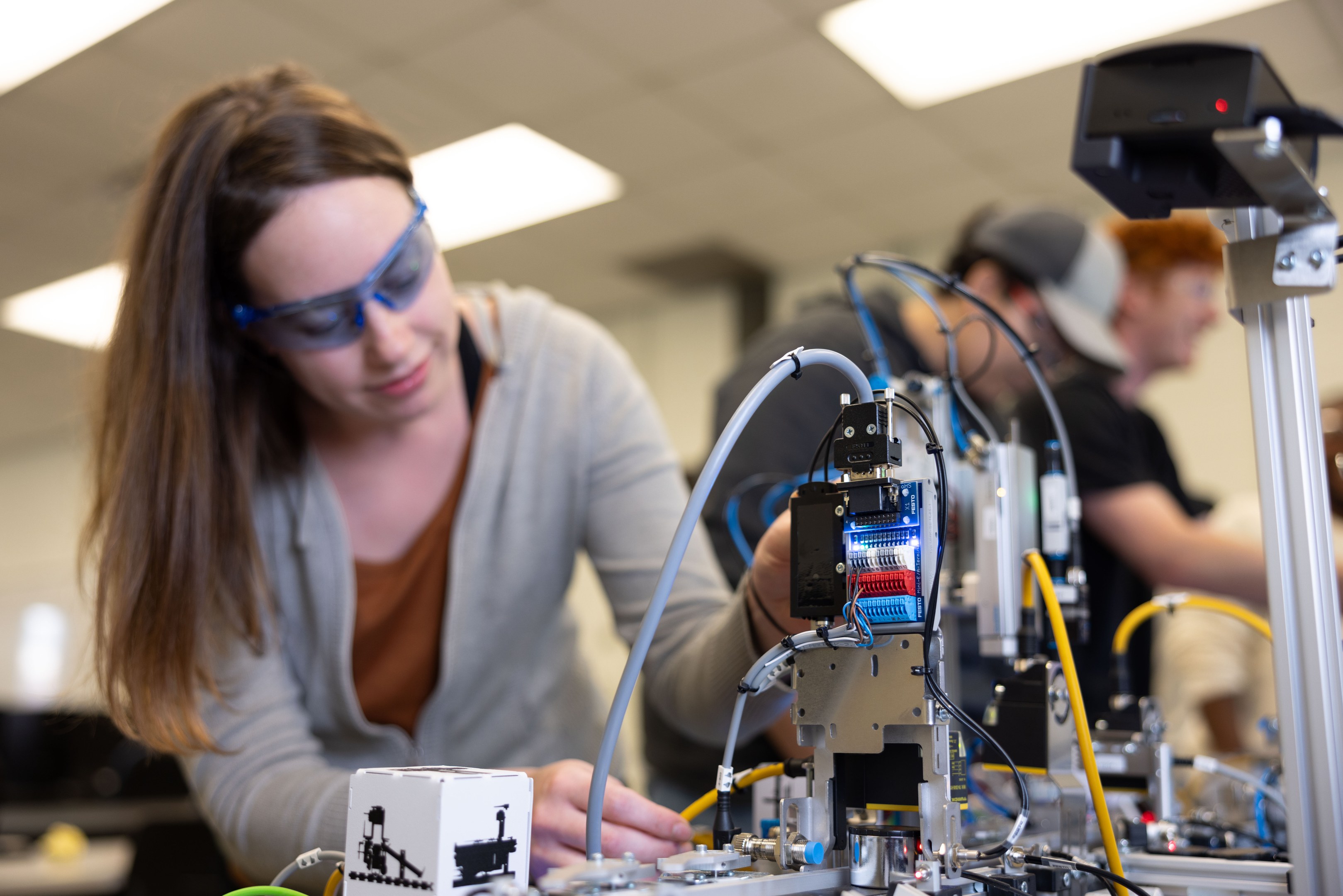Eine Frau mit Schutzbrille arbeitet an einem Roboter in einem Labor, umgeben von anderen, mit Werkzeugen und Bauteilen auf dem Tisch unter Deckenbeleuchtung.