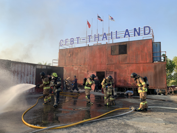 Feuerwehrleute in Helmen sprühen Wasser auf ein Gebäude mit Text, während Flaggen, Bäume und ein klarer blauer Himmel im Hintergrund zu sehen sind.