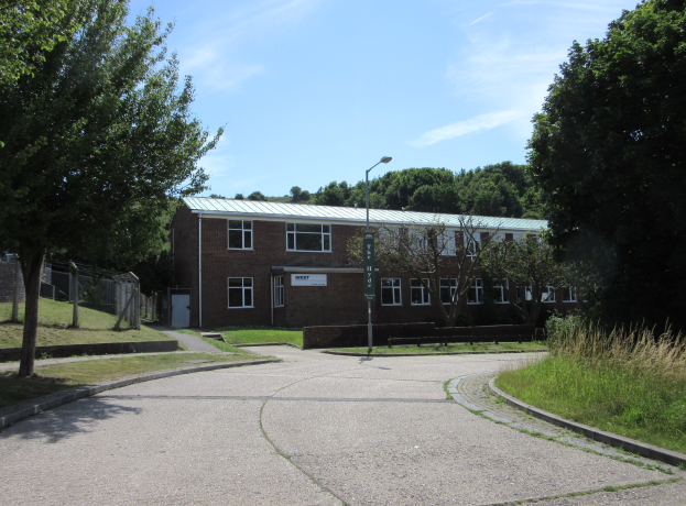 Ein Schulgebäude mit Fenstern, ein Straßenpfahl, ein Schild, Bäume, Gras, ein Zaun und ein bewölkter Himmel, umgeben von einem Park mit saftig grünem Gras.