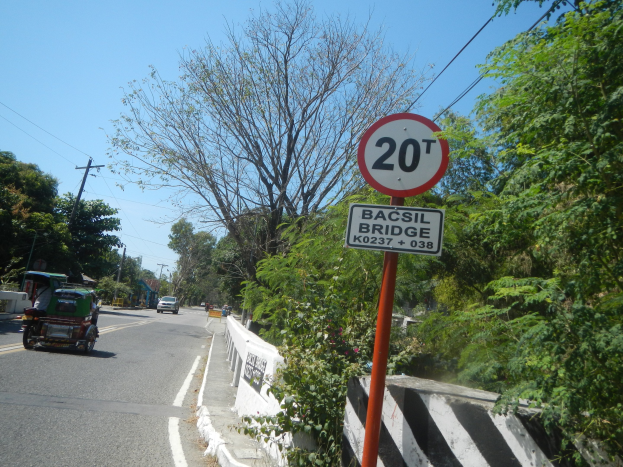 Eine geteilte Straße mit fahrenden Fahrzeugen, ein Tempolimit-Schild, Bäume, Strommasten mit Oberleitungen und einen klaren blauen Himmel.