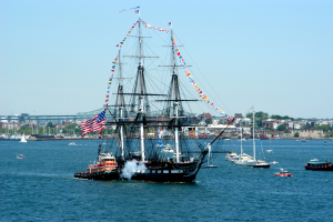 Ein Segelschiff mit Flaggen und Masten segelt auf dem Wasser in der Nähe einer Stadt, umgeben von anderen Booten, mit Gebäuden, Bäumen und einem klaren blauen Himmel im Hintergrund.