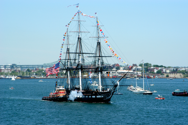 Ein Segelschiff mit Flaggen und Masten segelt auf dem Wasser in der Nähe einer Stadt, umgeben von anderen Booten, mit Gebäuden, Bäumen und einem klaren blauen Himmel im Hintergrund.