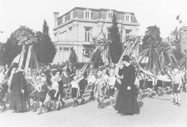 Gruppe von Menschen, die mit Fahnen vor einem Gebäude auf der Straße gehen, mit Hüten, Bäumen und Gebäuden im Hintergrund, in Schwarz-Weiß, mit einem Maibaum im Bild.