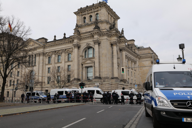 Eine Gruppe von Polizisten steht vor dem Reichstaggeb√§ude in Berlin, Deutschland, mit Fahrzeugen, einem Zaun, Verkehrszeichen, Laternenpf√§hlen, B√§umen und Flaggen im Hintergrund, bei klarem Himmel.