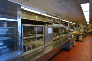 Gewerbeküche mit Edelstahl-Theke, aufgehängten Utensilien, einem Spülbecken, Armaturen, Schränken und diversen Gegenständen, die in Regalen unter elektrischem Licht angeordnet sind.