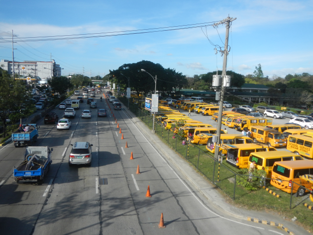 Eine belebte Straßenszene mit verschiedenen Fahrzeugen, Verkehrskegeln, Laternen, Strommasten mit Drähten und Texttafeln, mit einem Maschendrahtzaun auf der rechten Seite, Bäumen, Gebäuden und einem bewölkten Himmel im Hintergrund.