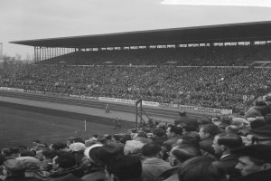 Schwarzes und weißes Foto eines vollen Stadions mit Zuschauern bei einem Fußballspiel, mit Bannern, Pfosten, einer Hütte, Bäumen, einem Turm und einem bewölkten Himmel.