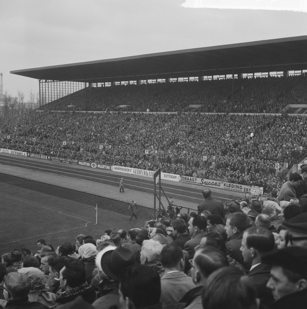 Schwarzes und weißes Foto eines vollen Stadions mit Zuschauern bei einem Fußballspiel, mit Bannern, Pfosten, einer Hütte, Bäumen, einem Turm und einem bewölkten Himmel.