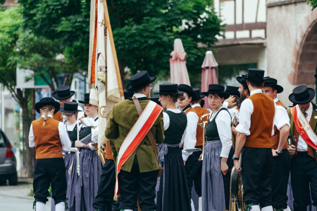 Gruppe von Menschen in traditioneller bayrischer Kleidung marschiert auf einer Straße, einige halten Musikinstrumente und Fahnen, mit Bäumen, Gebäuden und einem Auto im Hintergrund.