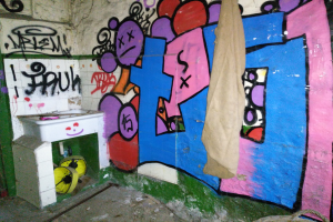 Ein mit Graffiti bedecktes Badezimmer mit einem in Stoff gehüllten Waschbecken und verstreuten Gegenständen auf dem Boden.