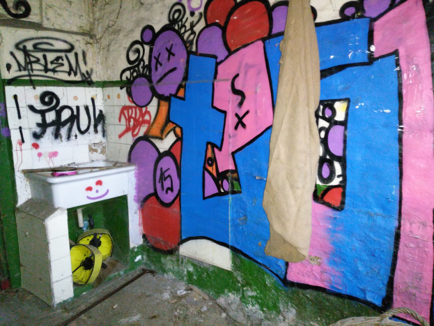 Ein mit Graffiti bedecktes Badezimmer mit einem in Stoff gehüllten Waschbecken und verstreuten Gegenständen auf dem Boden.